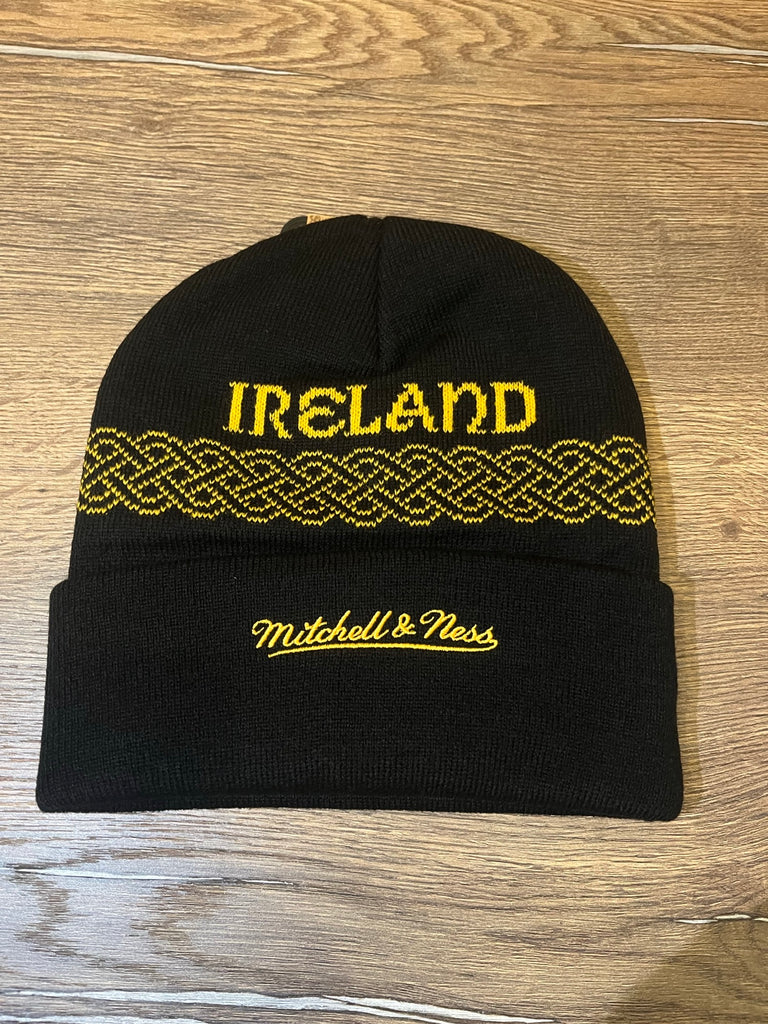 The Hat Knit Color Rush Ireland by Pittsburgh Steelers Europe Shop is a black beanie featuring yellow IRELAND text, a yellow Celtic knot pattern, and Mitchell & Ness embroidery on the cuff, shown on a wooden surface.