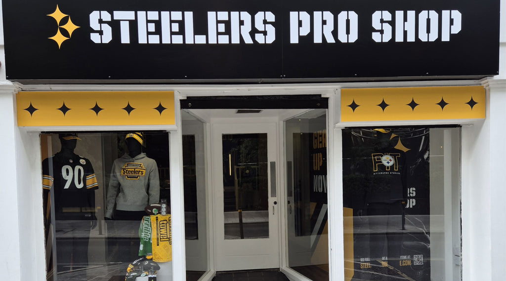 The storefront of a Steelers Pro Shop with black and gold signage, featuring Steelers jerseys, hats, and towels on display in the windows.