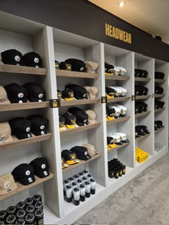 Shelves display various Pittsburgh Steelers hats in black, beige, and white, along with matching drinkware and tumblers. A sign above reads “HEADWEAR.” The items are neatly organized by color and style.