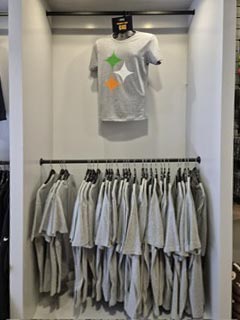 A display of gray t-shirts on hangers, with one t-shirt featuring a design of three colored stars (green, orange, and white) hanging above the others in a clothing store.
