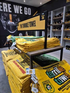 Stacks of yellow Terrible Towels are displayed on tables in a store, with shelves of hats and sports memorabilia in the background and a large football helmet image on the wall.